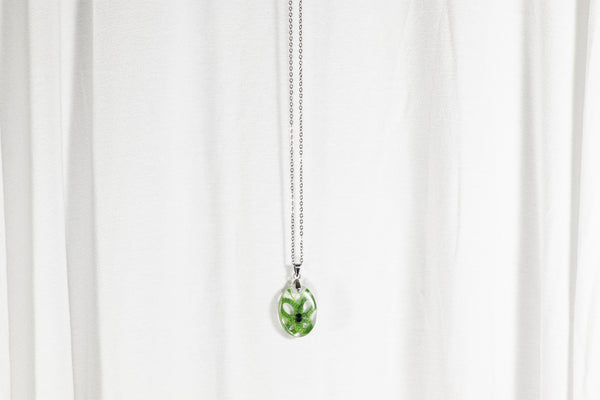 Majen ovalen obesek iz epoksidne smole z metuljem iz čipke v svetlo ali temno zeleni barvi. Visi na krajši verižici iz medenine oz. bakra.