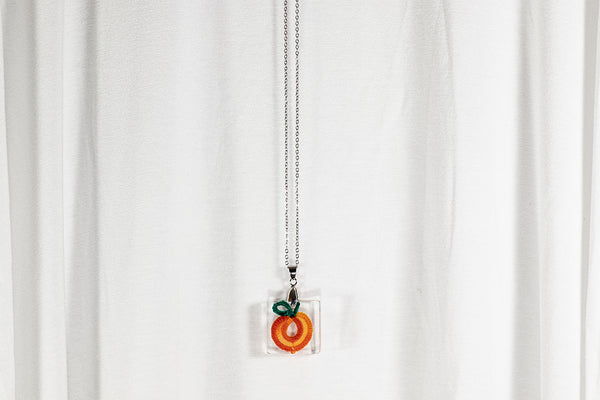 Manjši kvadraten obesek iz epoksidne smole z oranžno rumeno pomarančo iz čipke. Visi na krajši verižici iz medenine oz. bakra.