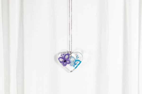 Obesek iz epoksidne smole v obliki srca, z vijoličnim in prelivajoče modrim metuljem iz čipke. Visi na verižici iz medenine oz. bakra.