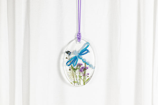 Čudovit večji ovalen obesek iz epoksidne smole, s prelivajoče modrim kačjim pastirjem iz čipke, ki leta na travniku vijolično modro belih sušenih cvetlic. Visi na vijolični vrvici iz povoščenega bombaža.