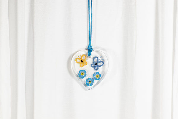 Obesek iz epoksidne smole srčaste oblike, z rumenim in modrim metuljem iz čipke ter tremi spominčicami iz polimerne gline. Visi na modri povoščeni bombažni vrvici.