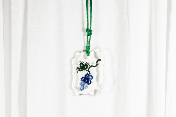 Obesek iz epoksidne smole vijugasto kvadratne oblike, na daljši temno zeleni vrvici iz povoščenega bombaža, s čipko v obliki prelivajoče se temno modrega grozda na temno zeleni vitici.