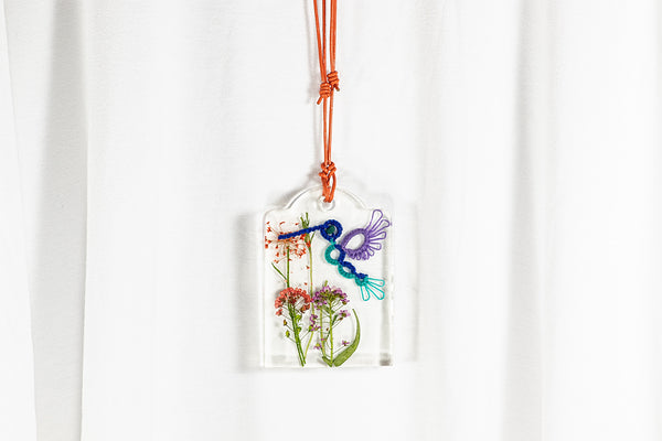 Obesek iz epoksidne smole na daljši vrvici iz opečnato rjavega usnja. V obesku je čipkast kolibri modro vijolične barve na rdeče vijoličnem sušenem cvetju.