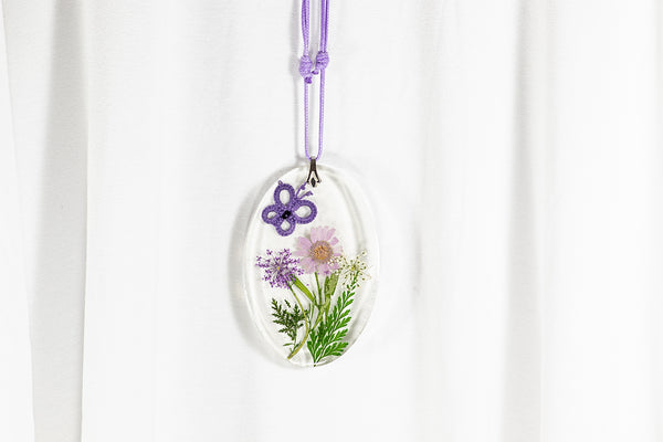 Večji ovalen obesek iz epoksidne smole z vijoličnim metuljem iz čipke na travniku vijolično belih sušenih cvetlic. Visi na vijolični povoščeni vrvici iz bombaža.
