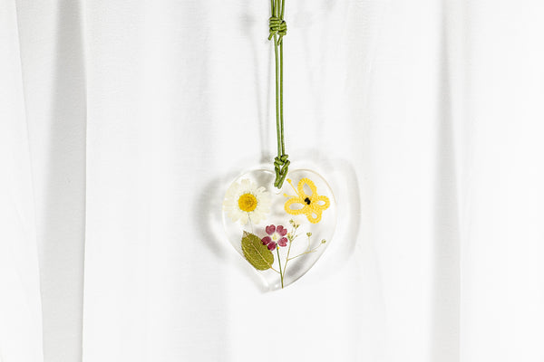 Obesek srčaste oblike iz epoksidne smole, z rumenim čipkastim metuljem in sušenim cvetjem rdeče in bele barve. Visi na usnjeni zeleni vrvici.