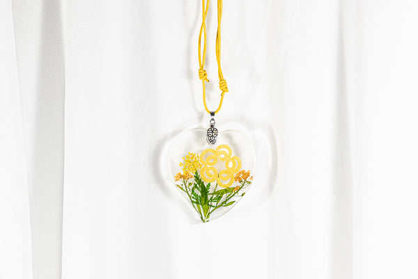 Srčast obesek iz epoksidne smole s cvetlico iz čipke in sušenim rumenim cvetjem. Visi na rumeni usnjeni vrvici.