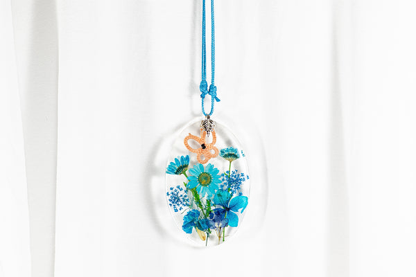 Večji ovalen obesek iz epoksidne smole z marelično oranžnim metuljem iz čipke na travniku turkizno modrih sušenih cvetlic. Visi na turkizno modri vrvici iz povoščenega bombaža.