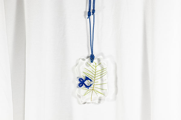 Obesek iz epoksidne smole v obliki vijugastega pravokotnika, na dalšji temno modri usnjeni vrvici. Z modro ribo narejeno iz čipke in belih perl, ter sušenim zelenjem.