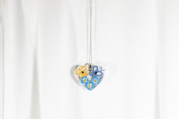 Obesek iz epoksidne smole v obliki srca, z rumenim in modrim čipkastim metuljem ter tremi spominčicami iz polimerne gline. Visi na verižici iz medenine oz. bakra.