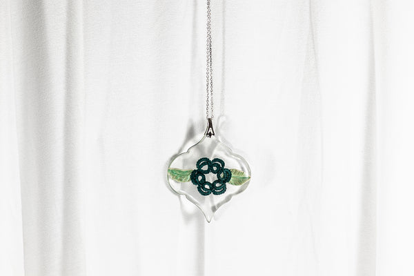 Obesek baročne oblike iz epoksidne smole, na verižici iz posrebrene medenine oz. bakra. S cvetom iz temno zelene čipke in dvema bledo zelenima listoma na vsaki strani.