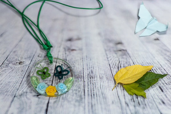 Okrogel obesek iz epoksidne smole, s svetlo in temno zelenim čipkastim metuljem, dvema svetlo modrima in eno rumeno vrtnico ter dvema bledo zelenima listoma na vsaki strani le-teh. Na zeleni povoščeni vrvici iz bombaža.