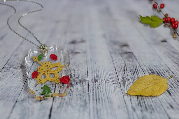 Obesek iz epoksidne smole, vijugasto pravokotne oblike, na dolgi verižici iz medenine oz. bakra. Z lepo rumeno čipko javorjevega lista, okrog so rdeči in zeleni listki iz polimerne gline.