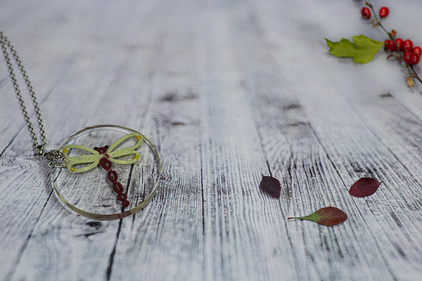 Večji okrogel obesek iz epoksidne smole, s čipkastim rdečim kačjim pastirjem z rumeno-zelenimi krili. Visi na verižici iz medenine oz. bakra.