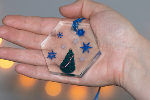 Šestkoten obesek iz epoksidne smole z decembrsko zgodbo: okrašena smrečica iz čipke z vijolično girlando, obdana z modrimi in belimi snežinkami iz polimerne smole. Na modri usnjeni vrvici.