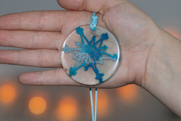 Okrogel obesek na svetlo modri bombažni vrvici iz epoksidne smole, s prelivajočo svetlo modro kvačkano čipko v obliki snežinke.