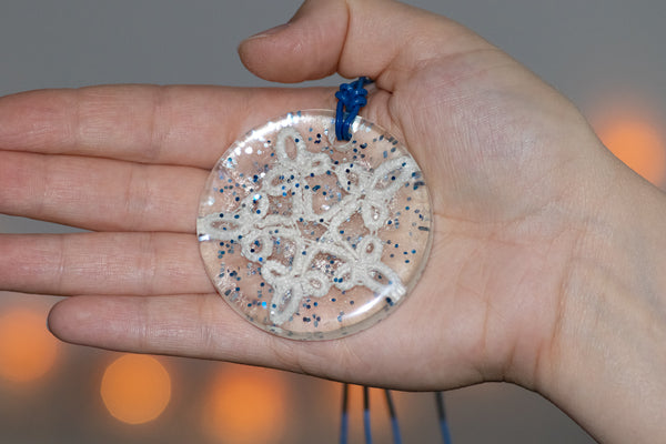 Večji okrogel obesek iz epoksiden smole z belo čipko v obliki zvezde ter modrimi bleščicami. Visi na temno modri usnjeni vrvici.