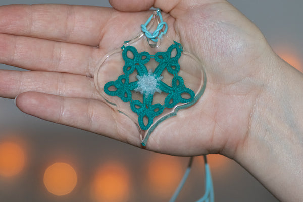 Obesek baročne oblike na dlani, ki je na svetlo modri usnjeni vrvici. Obesek je iz epoksidne smole in vsebuje zeleno modro čipko v obliki petkotne zvezde, ki ima majhno belo snežinko na sredini.