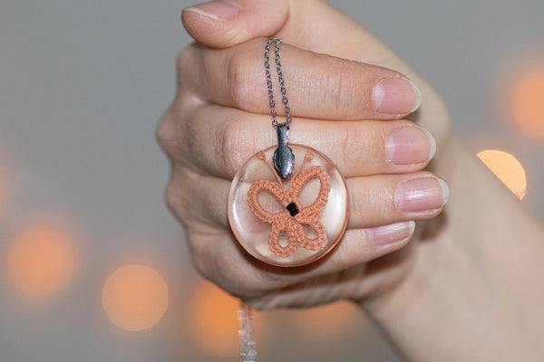 Okrogel obesek iz epoksidne smole z marelično oranžnim čipkastim metuljem. Visi na krajši verižici iz medenine oz. bakra.