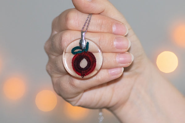 Okrogel obesek iz epoksidne smole z čipko v obliki temno rdečega sadja. Visi na verižici iz medenine oz. bakra.