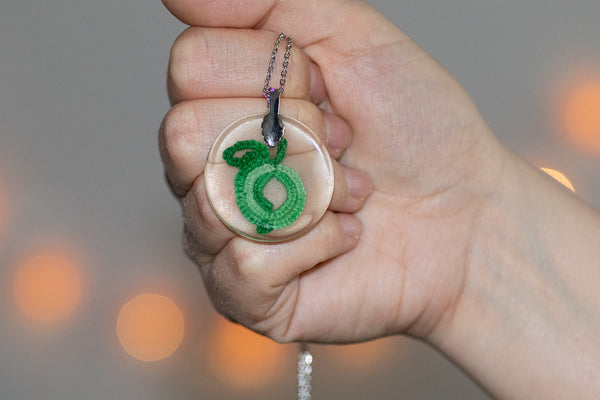 Manjši okrogel obesek iz epoksidne smole, z prelivajoče zelenim jabolkom iz čipke. Visi na krajši vrvici iz medenine oz. bakra.