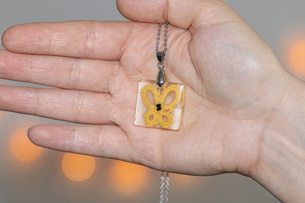 Manjši obesek kvadratne oblike iz epoksidne smole, z rumenim čipkastim metuljem. Visi na krajši verižici iz medenine oz. bakra.