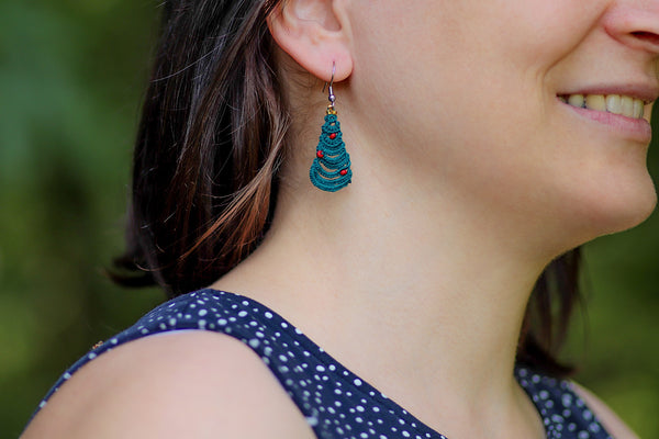 Čipkasti viseči uhani v obliki novoletne jelke. Na izbiro z rdečimi bunkicami ali zlato oz. vijolično girlando iz perlic. Visijo na kaveljčkih iz nerjavečega jekla.