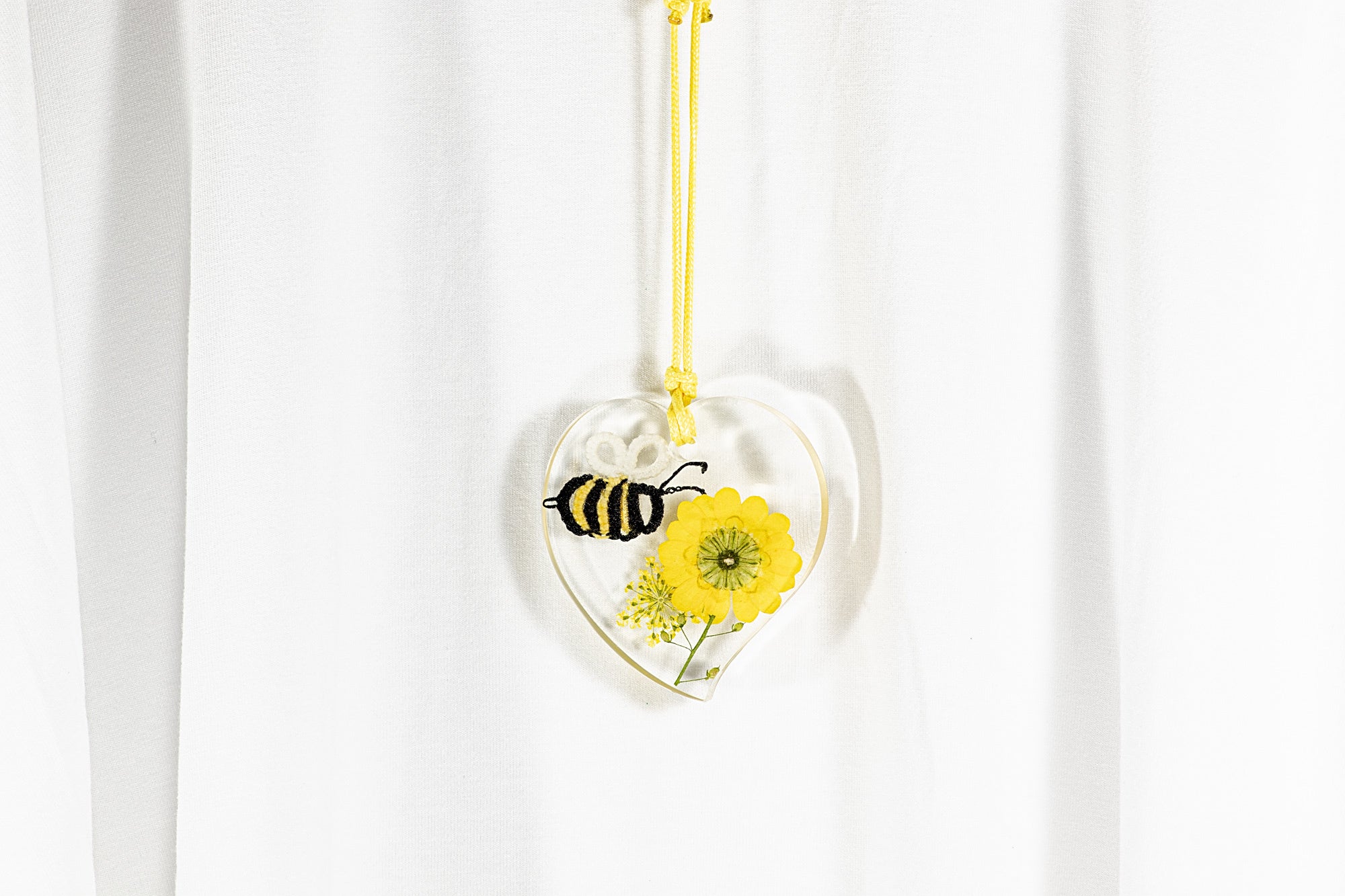 Obesek iz epoksidne smole v obliki srca, na daljši bombažni vrvici rumene barve, v katerem je čebela iz čipke na pravem sušenem rumenem cvetju.