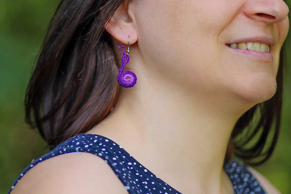 Čipkasti viseči uhani v obliki polžka prelivajoče vijolične barve, ki visijo na kaveljčkih iz nerjavečega jekla.